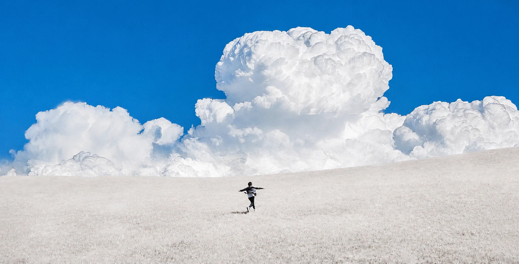 A small figure crossing a pale field beneath monumental white clouds and a blue sky